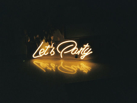 Neon sign reading "Let's Party" glows in warm yellow, casting a reflection on a surface in a dimly lit room, creating a festive mood.