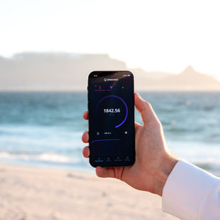 Test de vitesse 5G affiché sur un smartphone tenu à la main sur une plage, avec l’océan et des montagnes en arrière-plan.