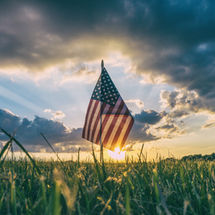 American flag in a grassy field at sunset, with rays of sunlight streaming through clouds, creating a serene and patriotic atmosphere.