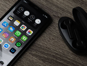 Smartphone displaying apps beside open black wireless earbuds on a wooden surface. Time on phone is 2:26. Background is neutral gray.