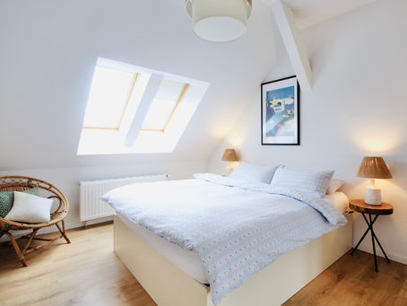Cozy attic bedroom with a skylight, double bed, blue patterned bedding, wicker chair, bedside lamps, and artwork, creating a serene mood.
