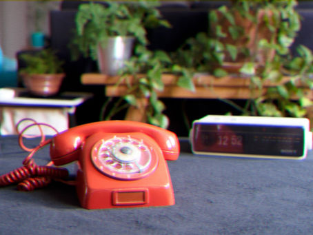 Red rotary phone on a table beside a digital clock showing 12:52. Background features green potted plants, creating a retro vibe.