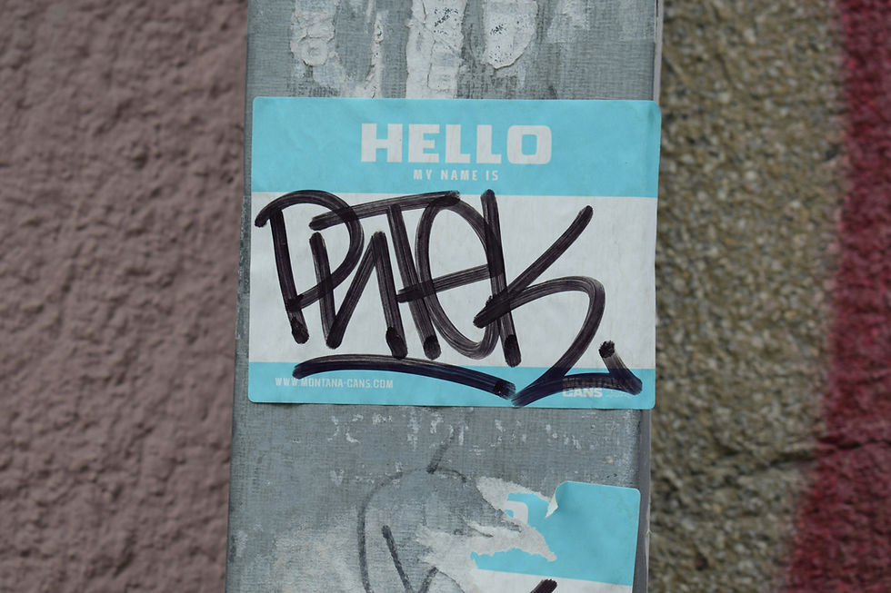 Blue HELLO MY NAME IS sticker on a weathered pole, covered with a black graffiti tag; textured wall in background.