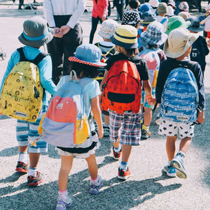 Children wearing colorful backpacks and hats walk outdoors, hand in hand, on a sunny day