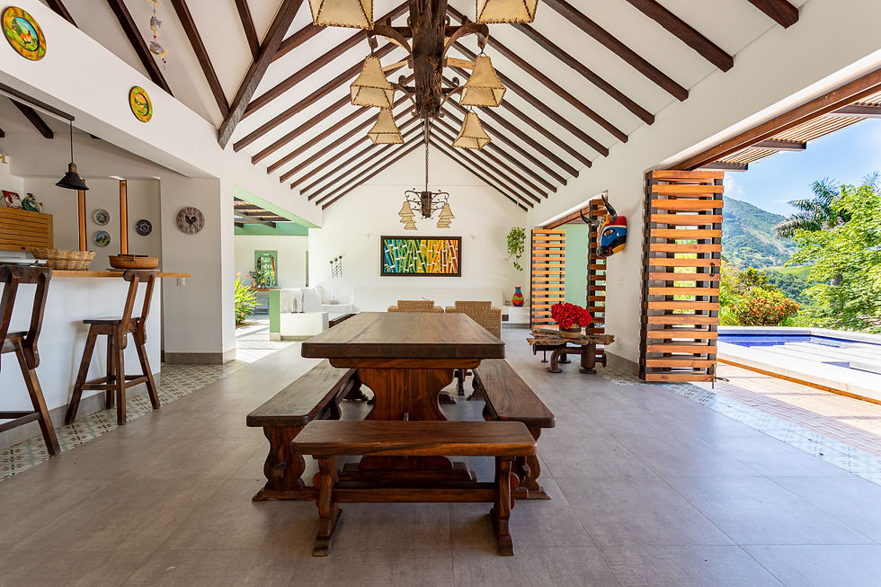 Spacious room with wooden beams, large table, and open doors to a scenic view. Bright colors, art on walls, and bar stools add charm.