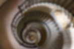 Spiral staircase view from above, showcasing black railing, checkered floor at the bottom, and brown marble walls, creating a dizzying effect.