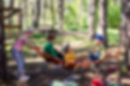 Four kindergarten aged children relax on a red rope hammock in a sunny pine forest, wearing colorful hats and shirts.