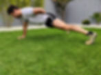 Man performing a one-arm push-up on green grass in a garden, wearing a gray shirt and black shorts, with a gray fence and plants behind.