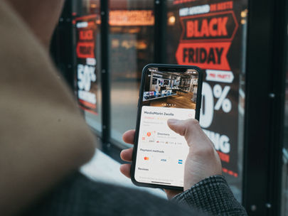 Person holds a phone displaying a store app near a shop window with bold Black Friday sale signs. Mood is focused and commercial.
