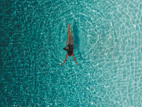 Person swimming in a clear turquoise pool, viewed from above. Ripples surround the figure, creating a tranquil, sunlit pattern.