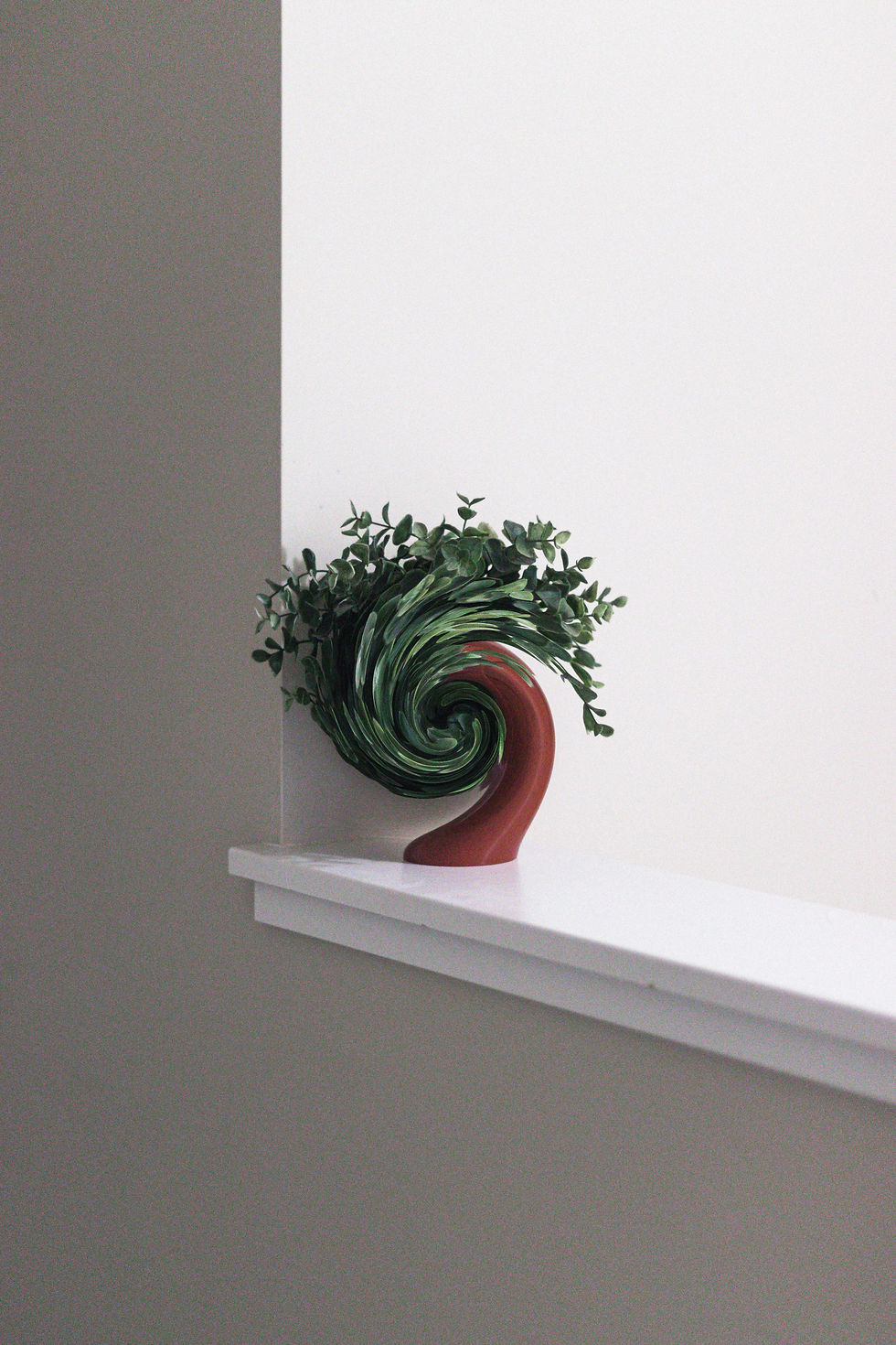 A plant in a red spiral-shaped vase sits on a white ledge against a gray and white wall, creating a minimalist aesthetic.