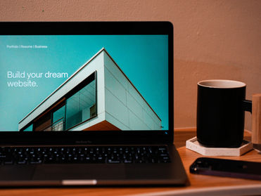 MacBook Pro on a wooden desk showing a website mockup with Build your dream website text, beside a black mug and phone.