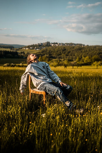 A casually dressed white young man leans back on a chair in the middle of a green summer meadow