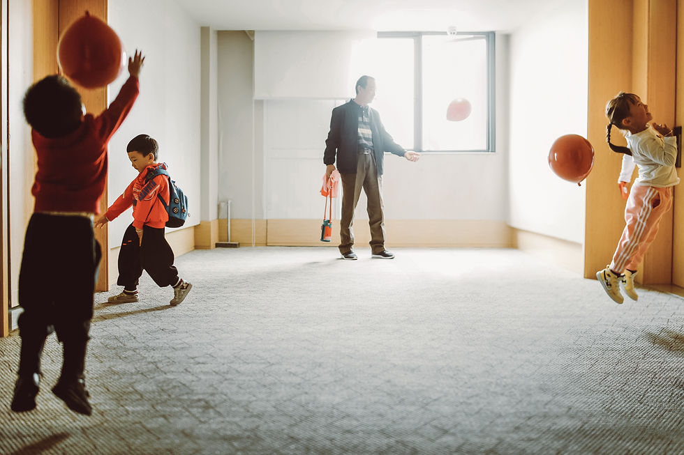 Children play with red balloons in a sunlit room. An adult watches, holding a toy. Joyful atmosphere with soft carpet and bright light.