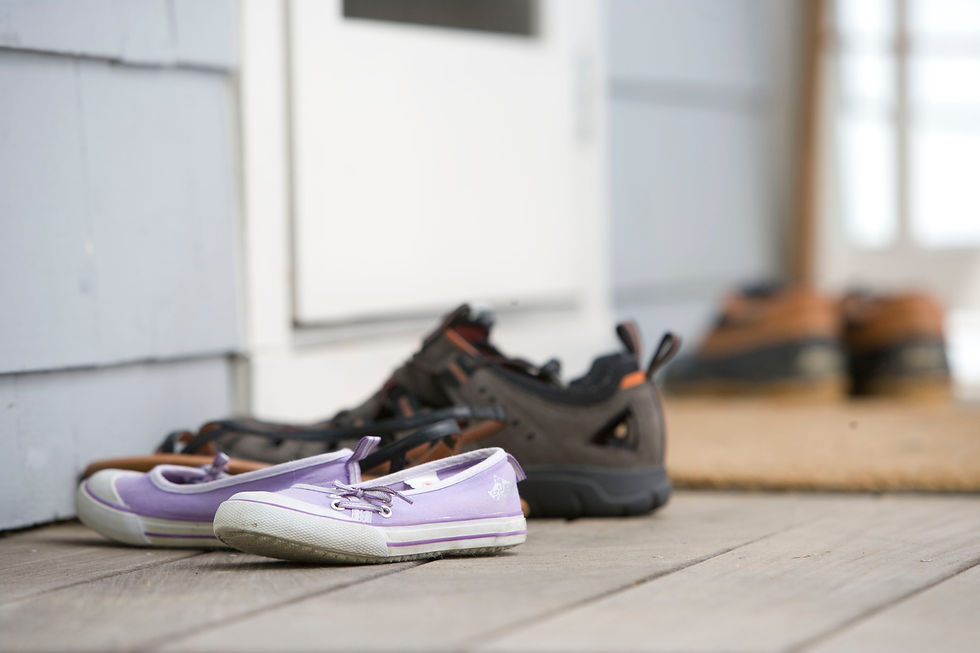 Purple sneakers and brown shoes on a wooden porch next to a blue house wall and a door, creating a cozy, welcoming atmosphere.