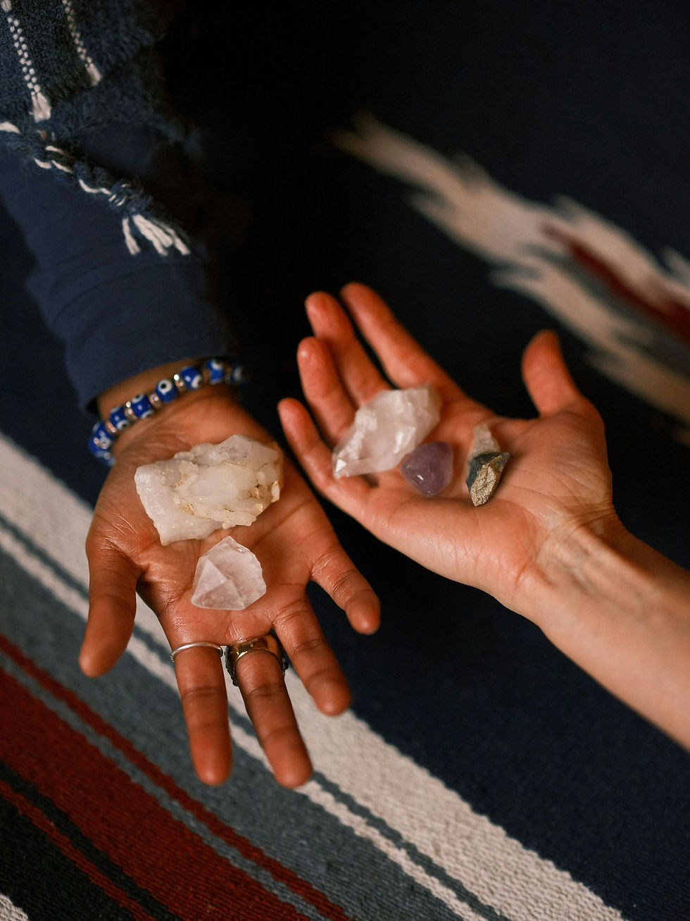 Two hands hold clear and purple crystals over a striped woven blanket, with rings and a beaded bracelet visible.