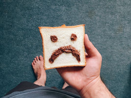Hand holds bread slice with chocolate spread frown and eyes on it. Bare feet visible on blue-grey floor. Playful mood.