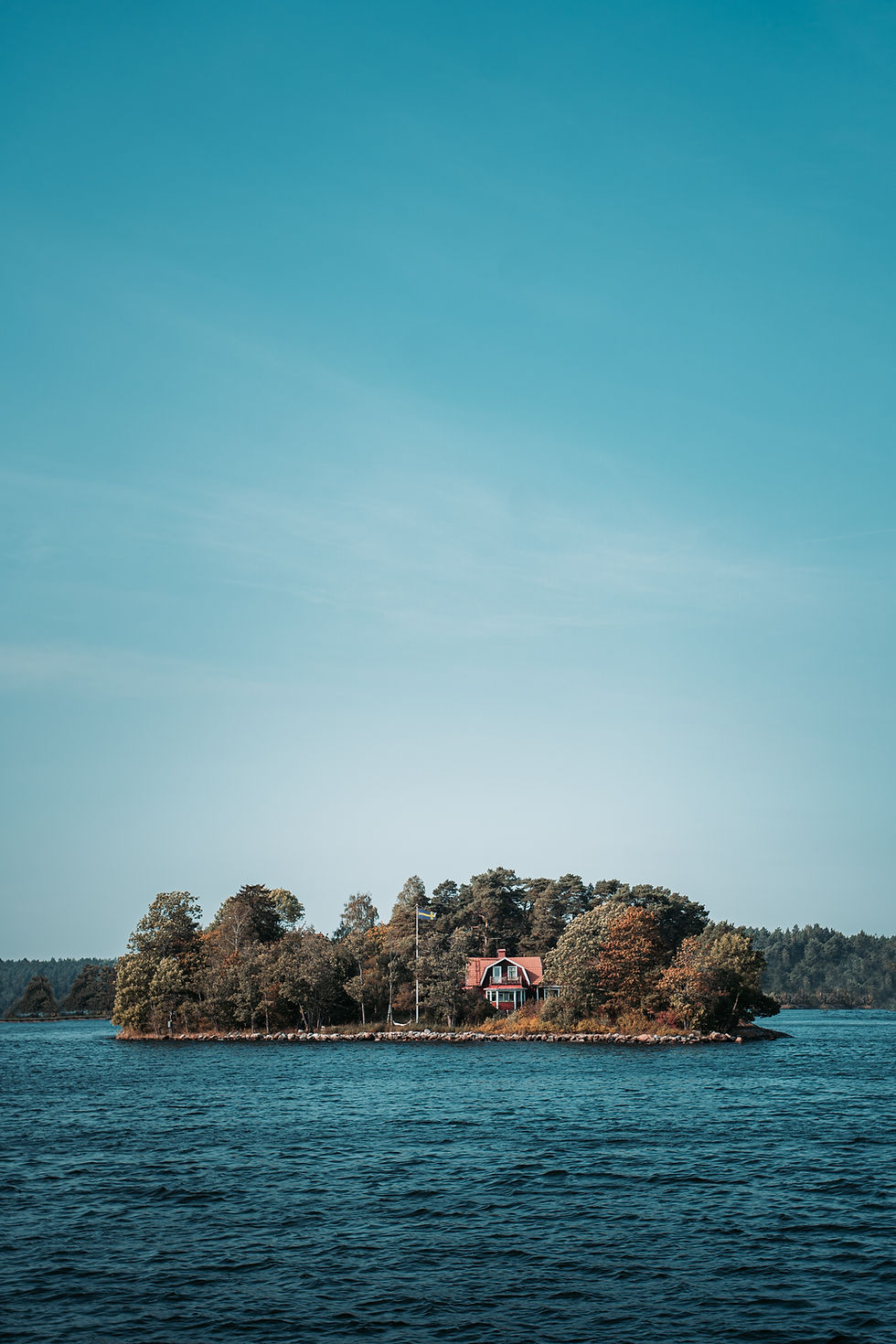 Petite île boisée avec maison rouge et drapeau suédois, entourée d’eau sous un vaste ciel bleu paisible.