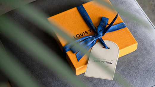 Orange gift box with blue ribbon and visible Louis Vuitton text. Set on a gray surface with blurred green leaves in the foreground.