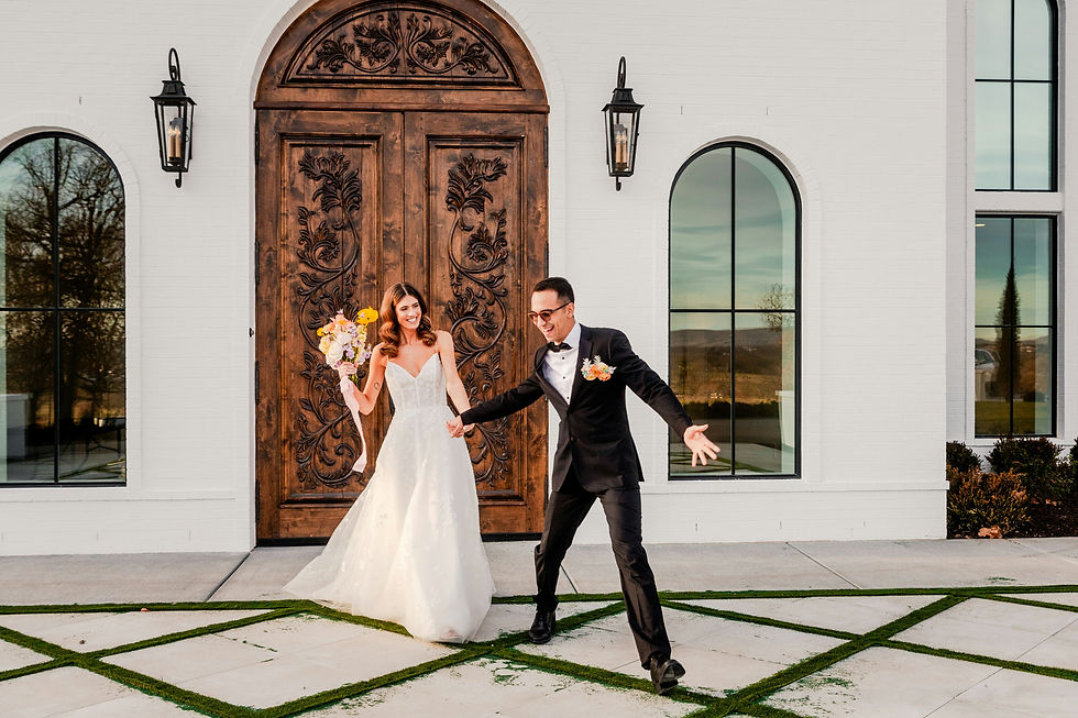 Bride in white gown holds bouquet, groom in black suit playfully gestures on stone path. Ornate wooden door, white building backdrop.