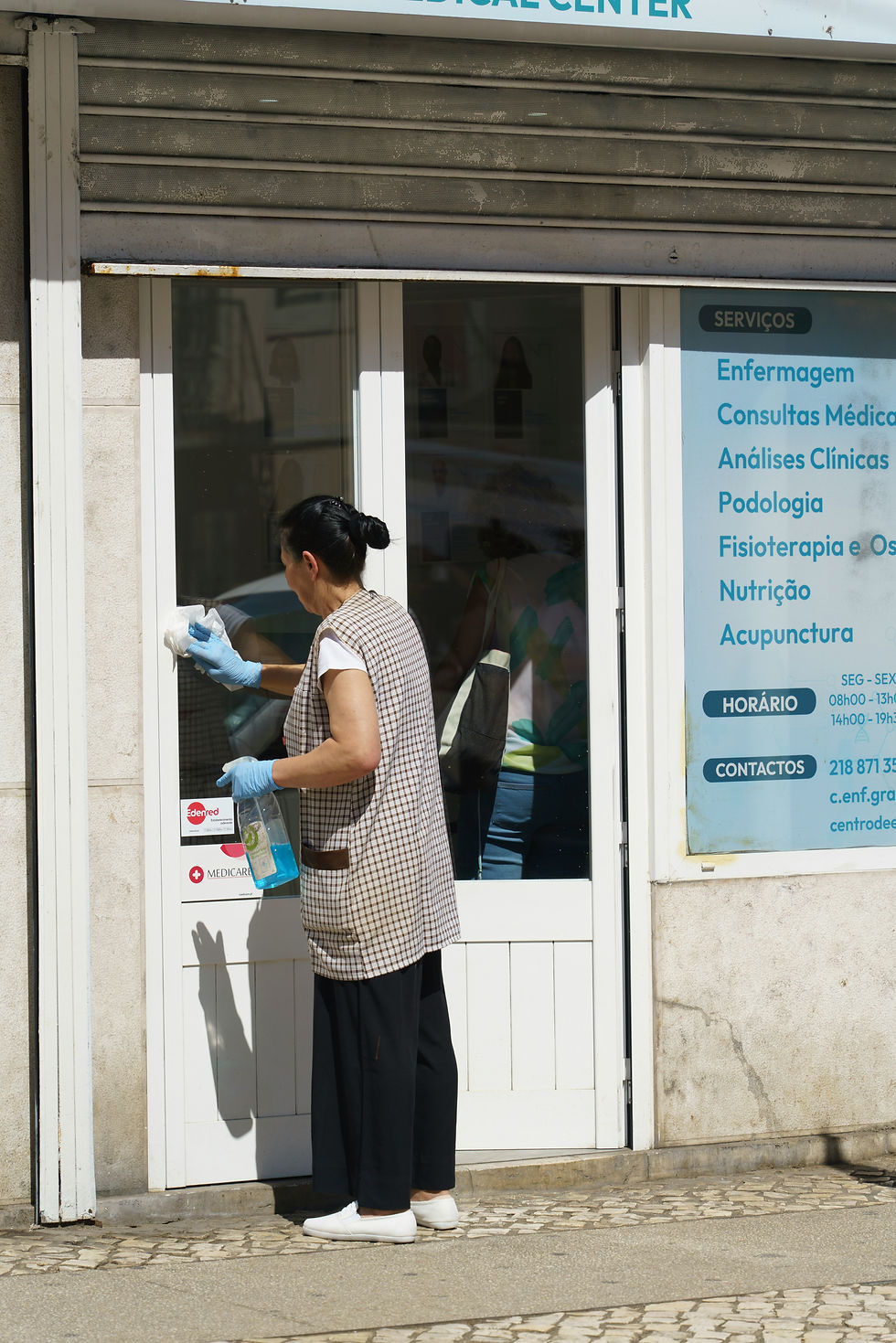 A woman cleans a glass door of a medical center with a spray and cloth. She's wearing gloves. Sunlit exterior with text listing services.