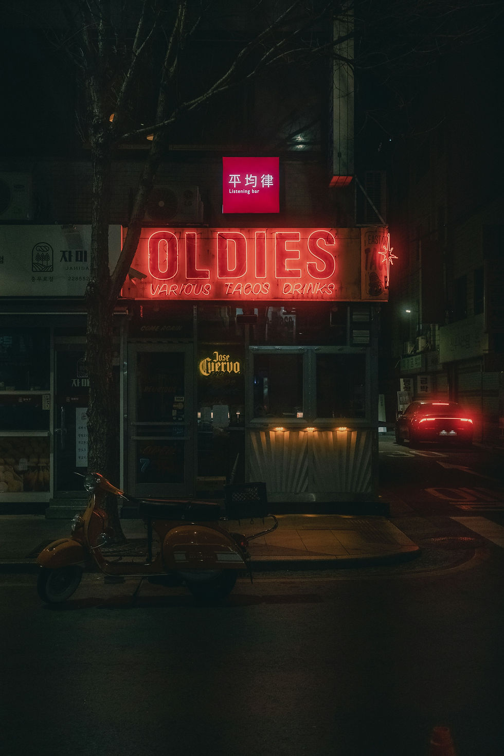 Neon signs reading "OLDIES" and "Listening Bar" glow in a dim city street. An orange scooter is parked in front, and a car passes by.