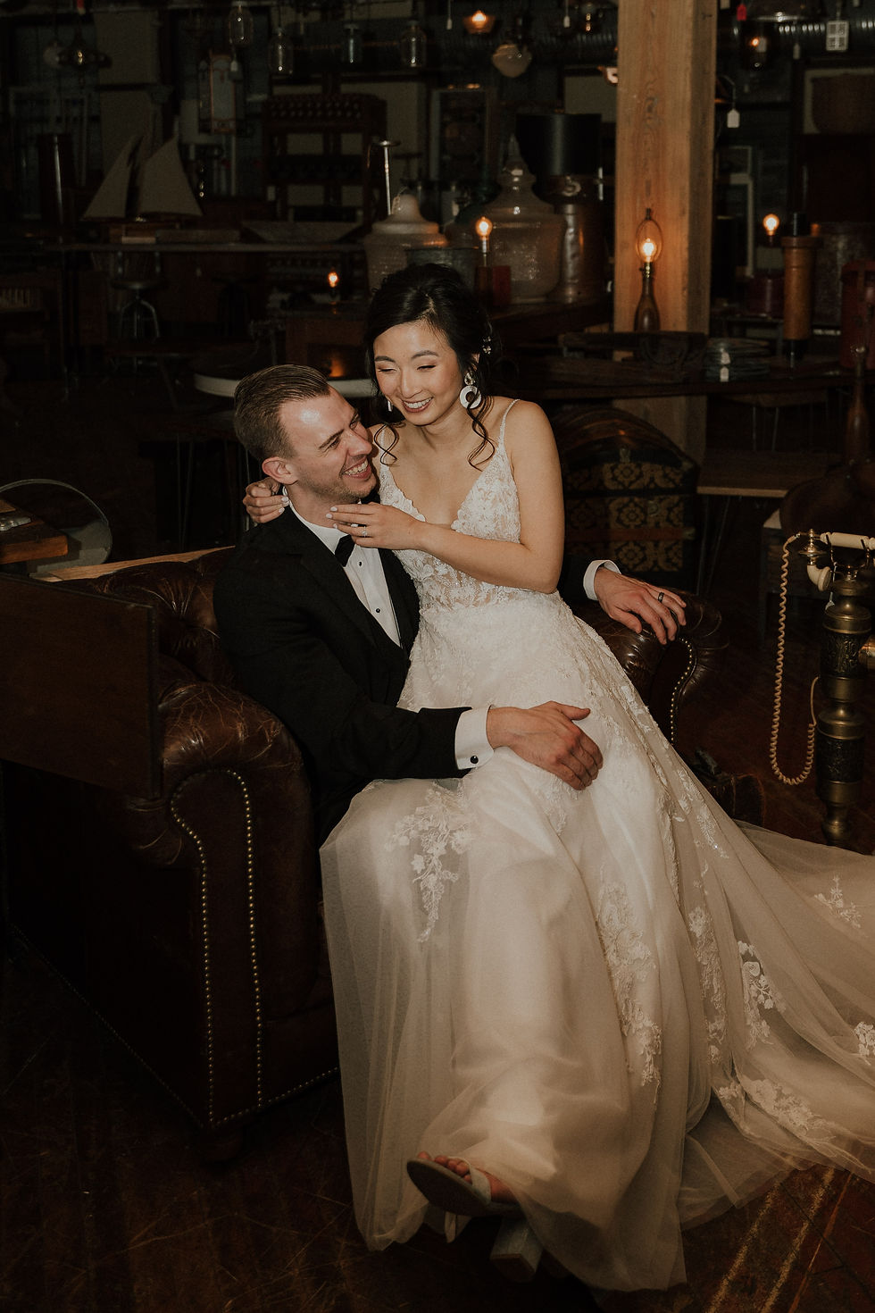 A joyful couple shares a tender moment, elegantly dressed for their wedding reception in a vintage, warmly lit room.