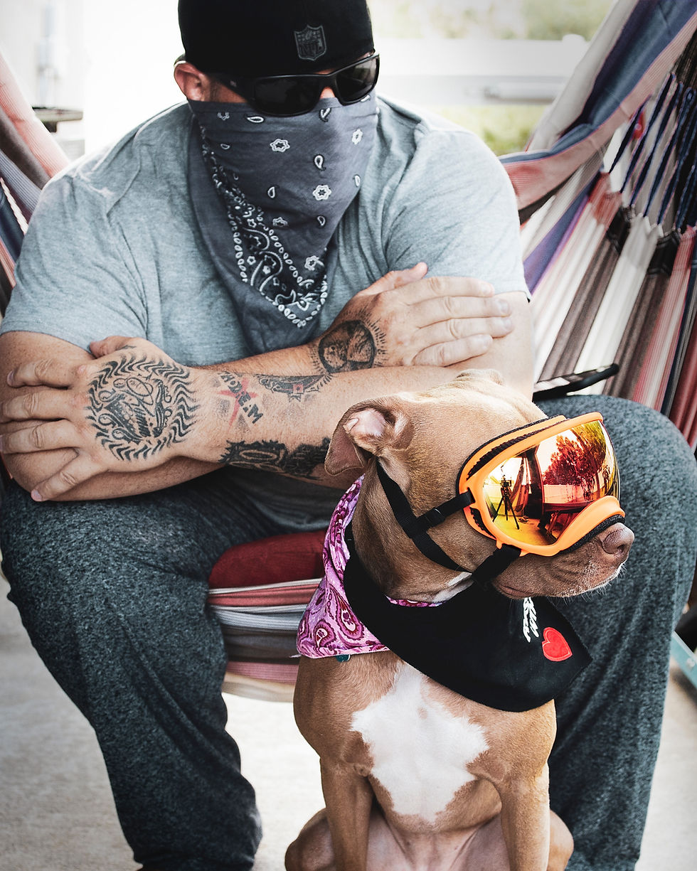 “A tattooed man sitting outdoors with his dog wearing reflective goggles and a bandana — a stylish, modern dog dad with his adventurous pup.”