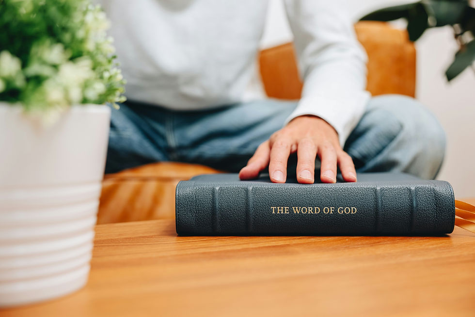A person sits on a couch, hand resting on a black book titled "THE WORD OF GOD" on a wooden table. A potted plant is nearby.