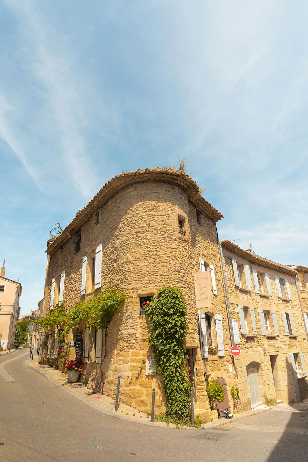 Charming stone building on a sunny street corner with ivy and red flowers. Blue sky above. Sign and stop symbol visible. Peaceful ambiance.
