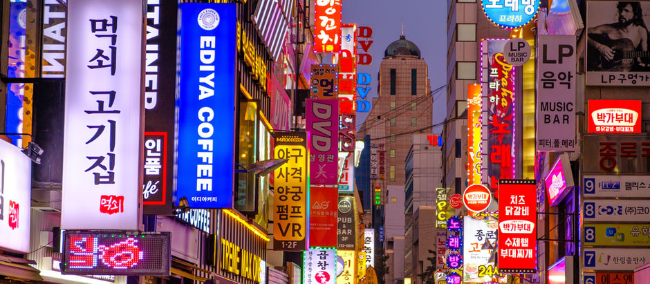 Neon-lit Korean nightlife street packed with tall shop signs for coffee, music bars, DVD and food under a dusk sky.