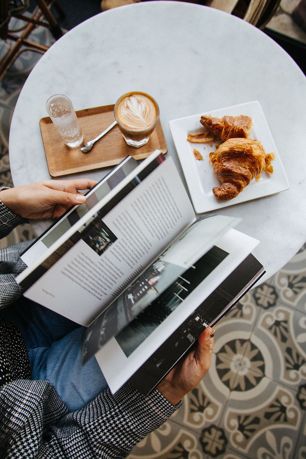 A woman reading a book at a table with a coffee and croissant