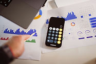 Smartphone com calculadora sobre relatórios com gráficos azuis; mão aponta para um gráfico em mesa de trabalho.