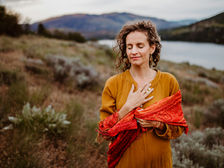 Frau mit ockerfarbener bluse, orangenem Schal und locken, steht in der Natur bei einem Fluss, hat die Augen geschlossen und hält sich ihre rechte Hand aufs Herz.