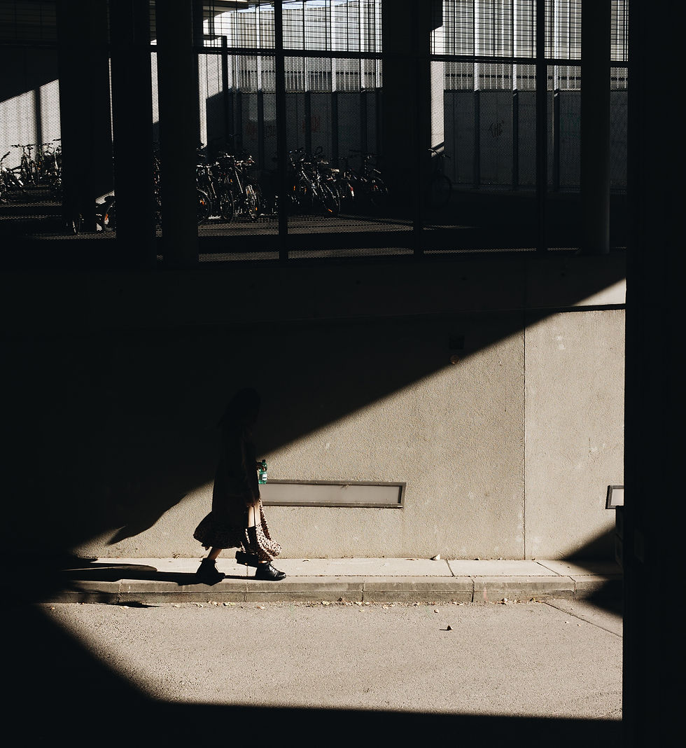 A person walks in shadow carrying a bag, with bicycles parked behind a fence. Sunlight creates geometric patterns on the concrete wall.