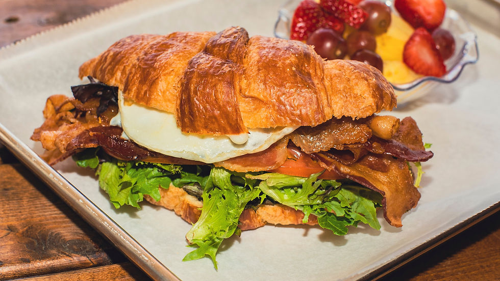Croissant sandwich with egg, bacon and lettuce on a tray, with a bowl of fruit and berries in the background.