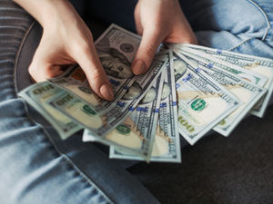 Hands hold fanned-out 100 dollar bills over blue jeans. Gray background. Text shows Benjamin Franklin's portrait. Mood: secure.