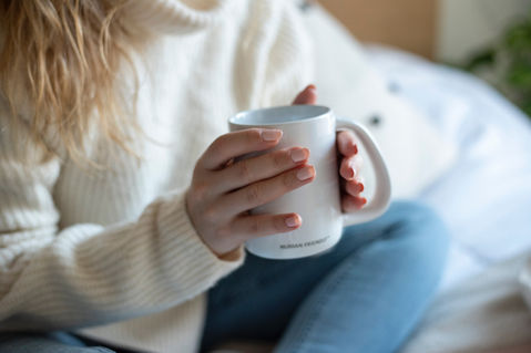 Close-up of a woman in a cream-colored sweater and blue jeans holding a white mug with both hands.