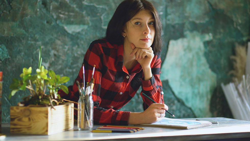 Young woman in a red plaid shirt sketches at a desk with art supplies, looking thoughtful against a green textured wall.
