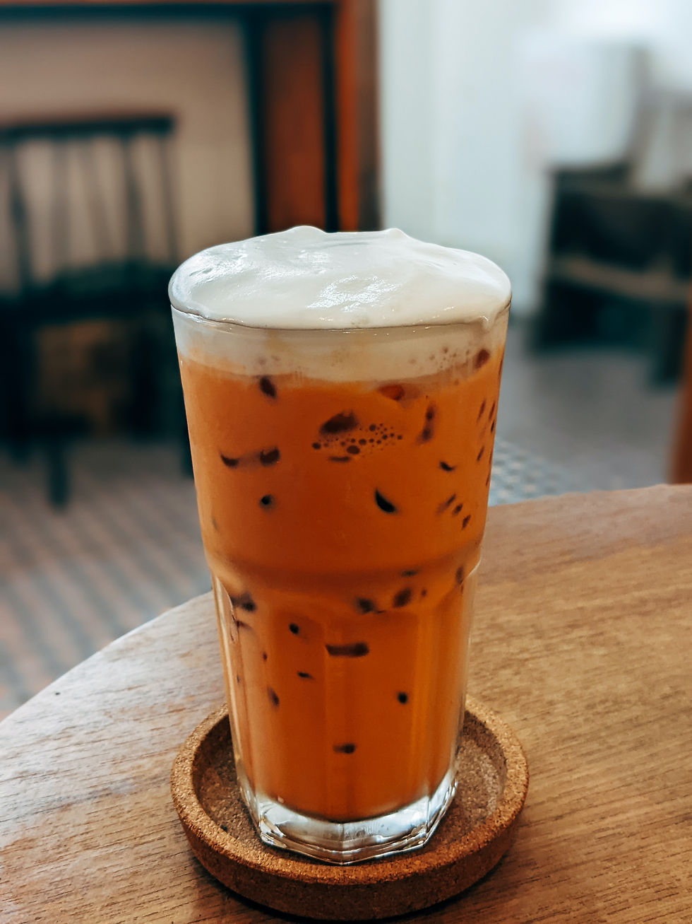 Iced Thai tea with creamy foam in a glass on a wooden table, with a cozy, blurred cafe background. Warm and inviting ambiance.