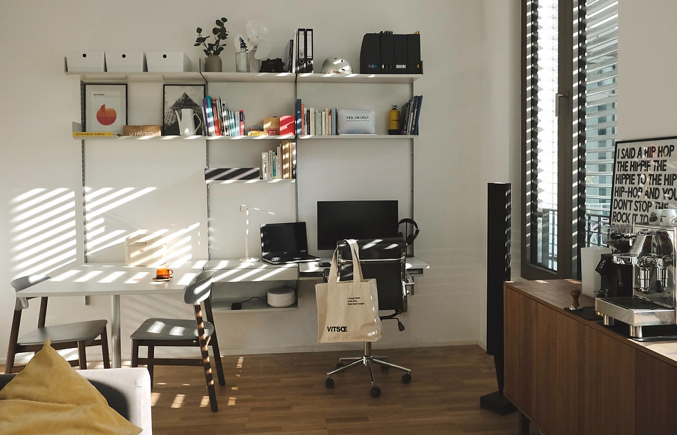 Home office with shelves, books, and decor. Sunlight casts striped shadows. Desk with laptop and tote bag. Modern and organized space.