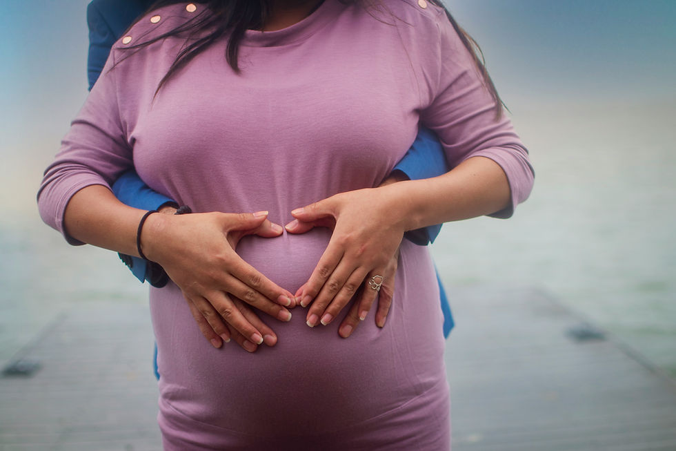Pregnant woman in a pink dress, hands forming a heart on her belly. Another person's arms embrace her from behind. Serene outdoor setting.