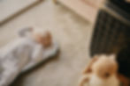 Baby in a white onesie sleeps on a mat on beige carpet. A plush toy and black suitcase nearby. Peaceful, warm setting.