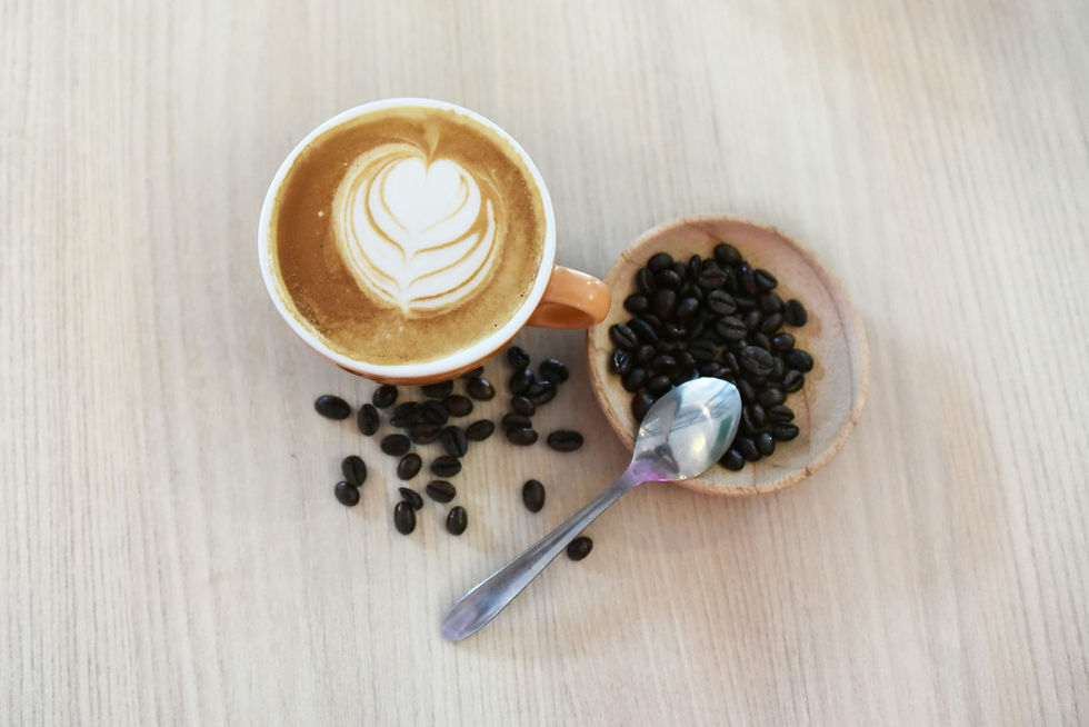 A cup of coffee with heart-shaped latte art sits beside a small bowl of coffee beans and a spoon on a light wooden surface, creating a cozy coffee scene.