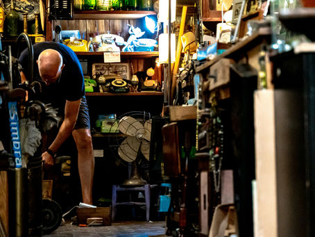 Bald man in a vintage shop bends over near antiques. Shelves display phones, fan, and signs. Warm lighting creates a nostalgic atmosphere.