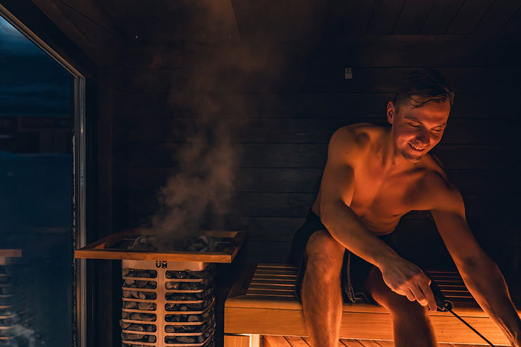 Man smiling in a traditional Finnish sauna with steam rising from a wood-burning heater