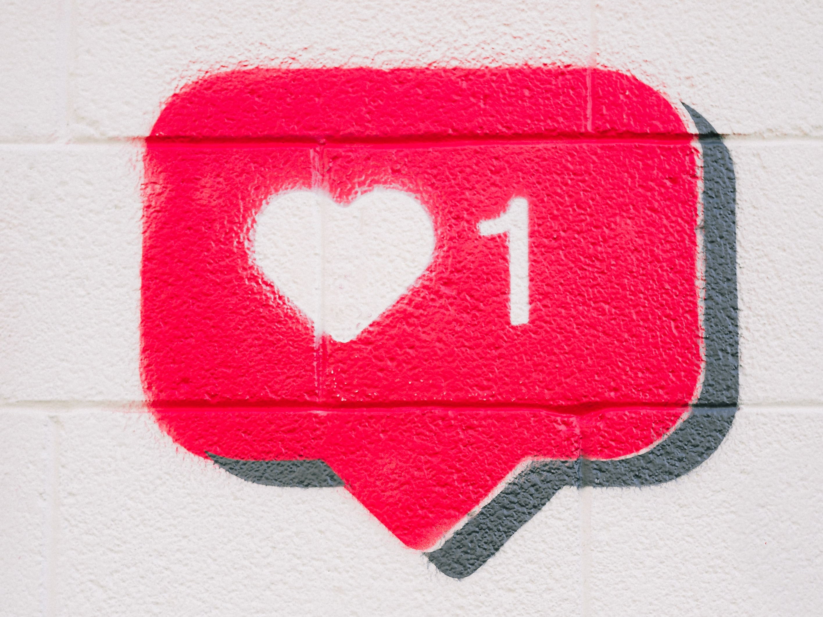 Hand holding a smartphone toward a wall mural of a pink heart notification with 1 like.