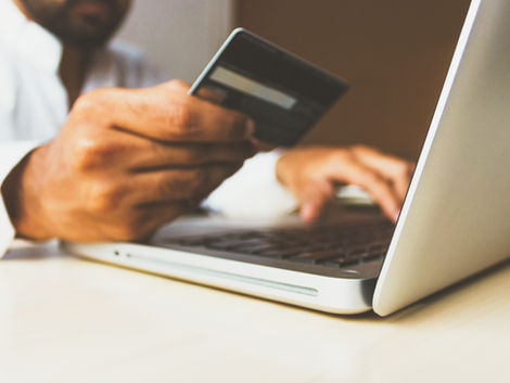 Person holding a credit card in front of a laptop on a wooden desk, indicating online shopping or payment. Muted colors, focused mood.