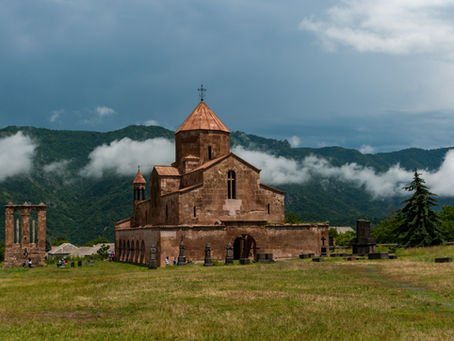 Stone church with copper roof in a grassy field, backed by misty green mountains and clouds; a few visitors nearby.