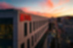 Netflix building with prominent red logo at sunset. City street below, lined with palm trees and lit by streetlights, under a pink-orange sky.
